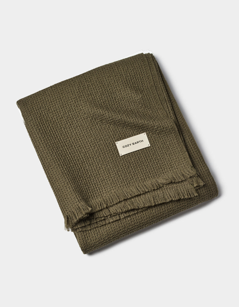 A folded olive green Alpaca Waffle Throw by Cozy Earth features a waffle texture, fringed edges, and a white tag with the brand name, set against a plain white background.