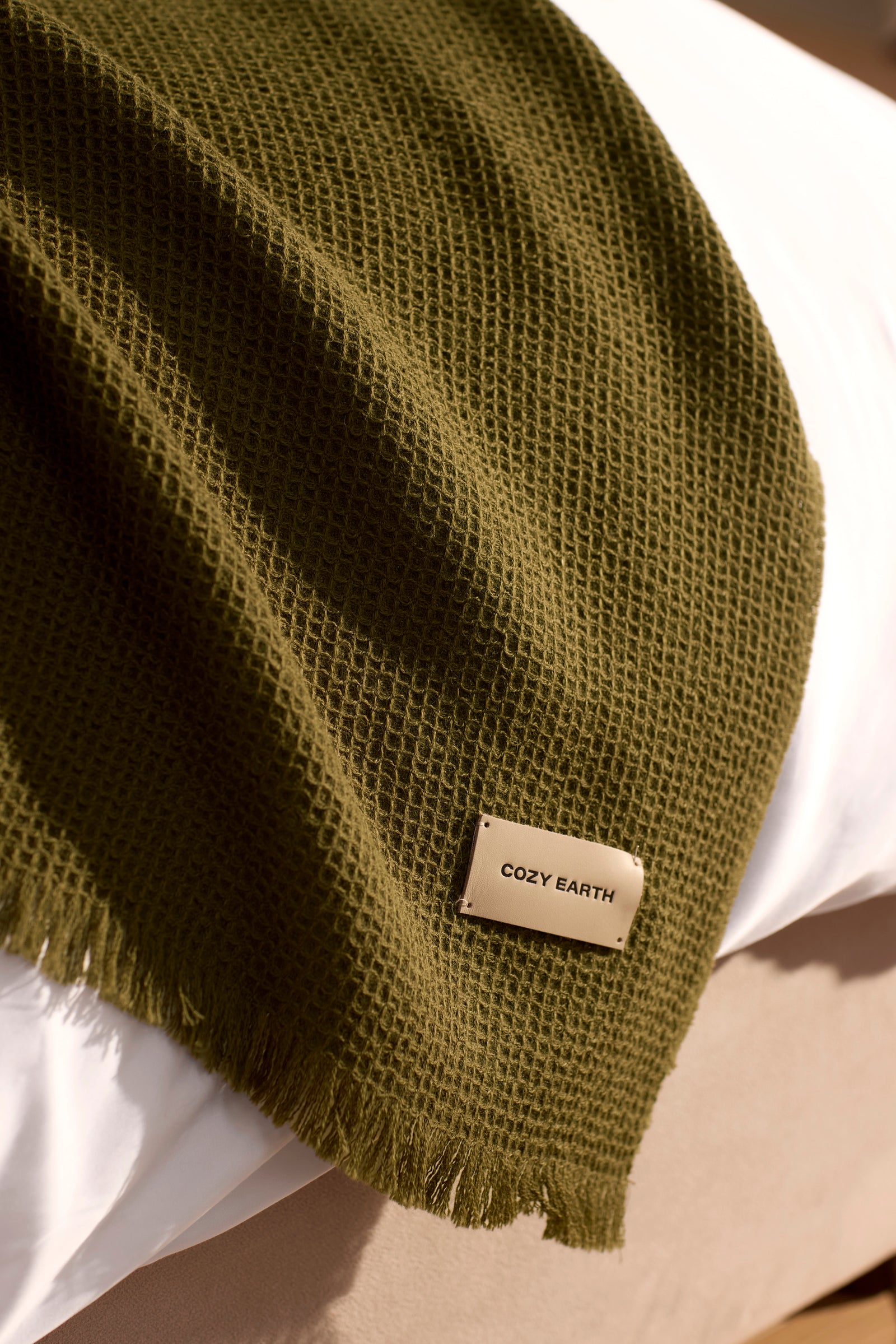 A Cozy Earth Alpaca Waffle Throw in textured olive green with fringe edges is draped over a white bed. A rectangular tag on the blanket displays "COZY EARTH" in black letters.