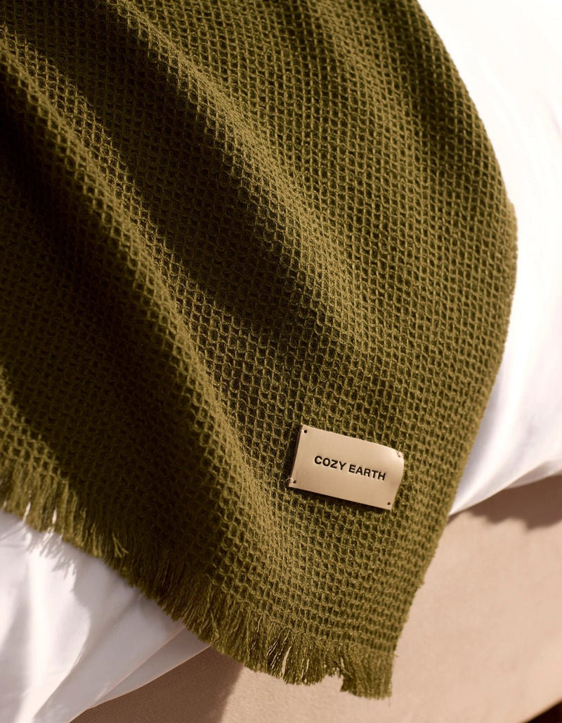 A Cozy Earth Alpaca Waffle Throw in dark green, featuring a textured waffle weave and fringed edges, is draped over a white surface. A beige tag on the throw displays the brand name "Cozy Earth.