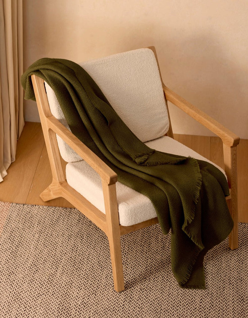 A wooden armchair with cream-colored cushions sits on a woven rug. A Cozy Earth Alpaca Waffle Throw in dark green is draped over the chair, adding a cozy and inviting touch.