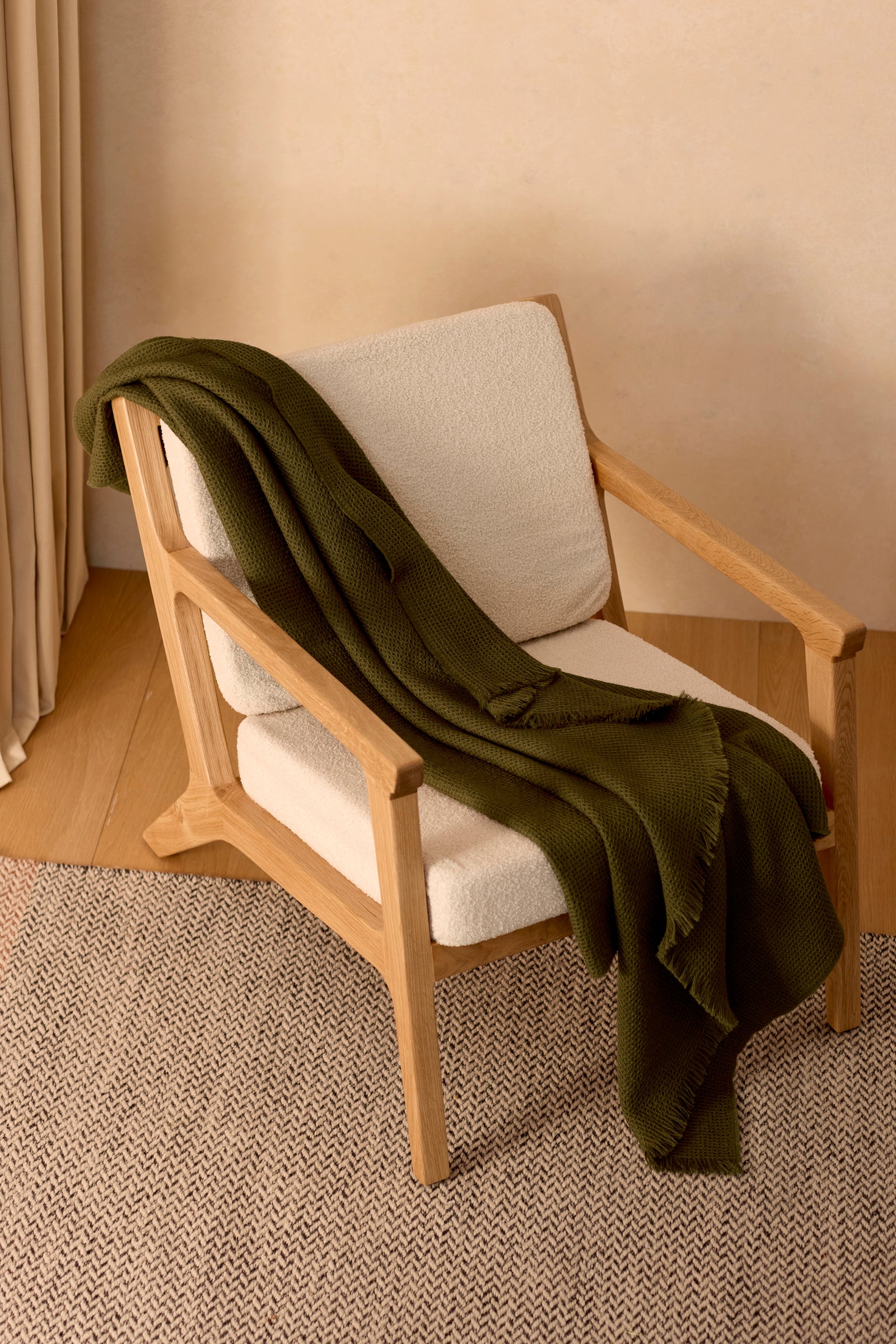 A modern wooden armchair with white cushions features a Cozy Earth Alpaca Waffle Throw in olive green draped over one arm. The chair rests on a textured rug in a softly lit room with beige walls and light wood flooring.
