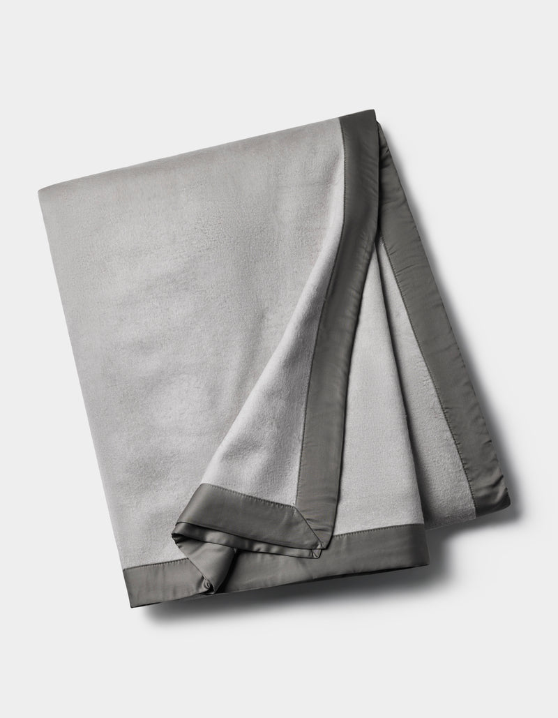 The Cozy Earth Bamboo Blanket is a folded light gray fleece with a smooth, dark gray satin border, shown on a plain white background.