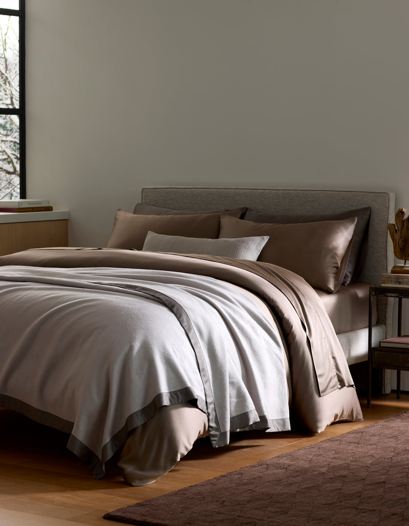 A neatly made bed features the Cozy Earth Bamboo Blanket in brown and light gray, with pillows against a gray upholstered headboard beside a window and wooden nightstand on hardwood floors.