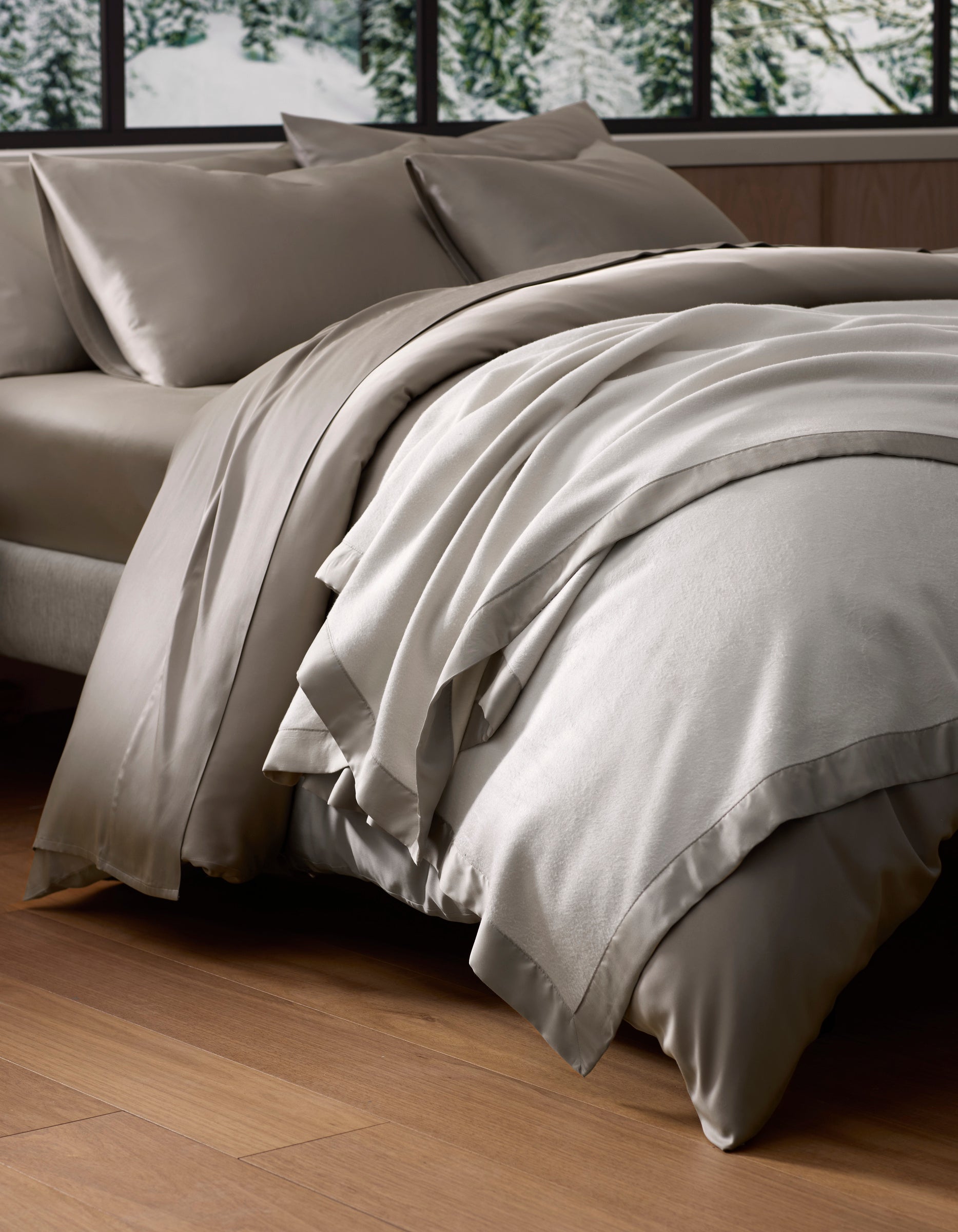 A neatly made bed with a luxurious Cozy Earth Bamboo Blanket in light gray, set near large windows overlooking snowy trees, on wooden floors in a cozy, modern room.|Color:Oat/Driftwood