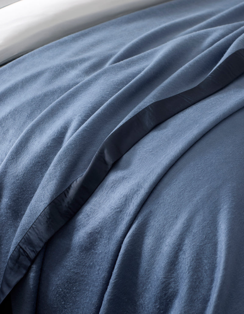 A close-up of a bed featuring the Cozy Earth Bamboo Blanket, showcasing its soft blue velvet-like texture, smooth folds, and a satin edge trim.