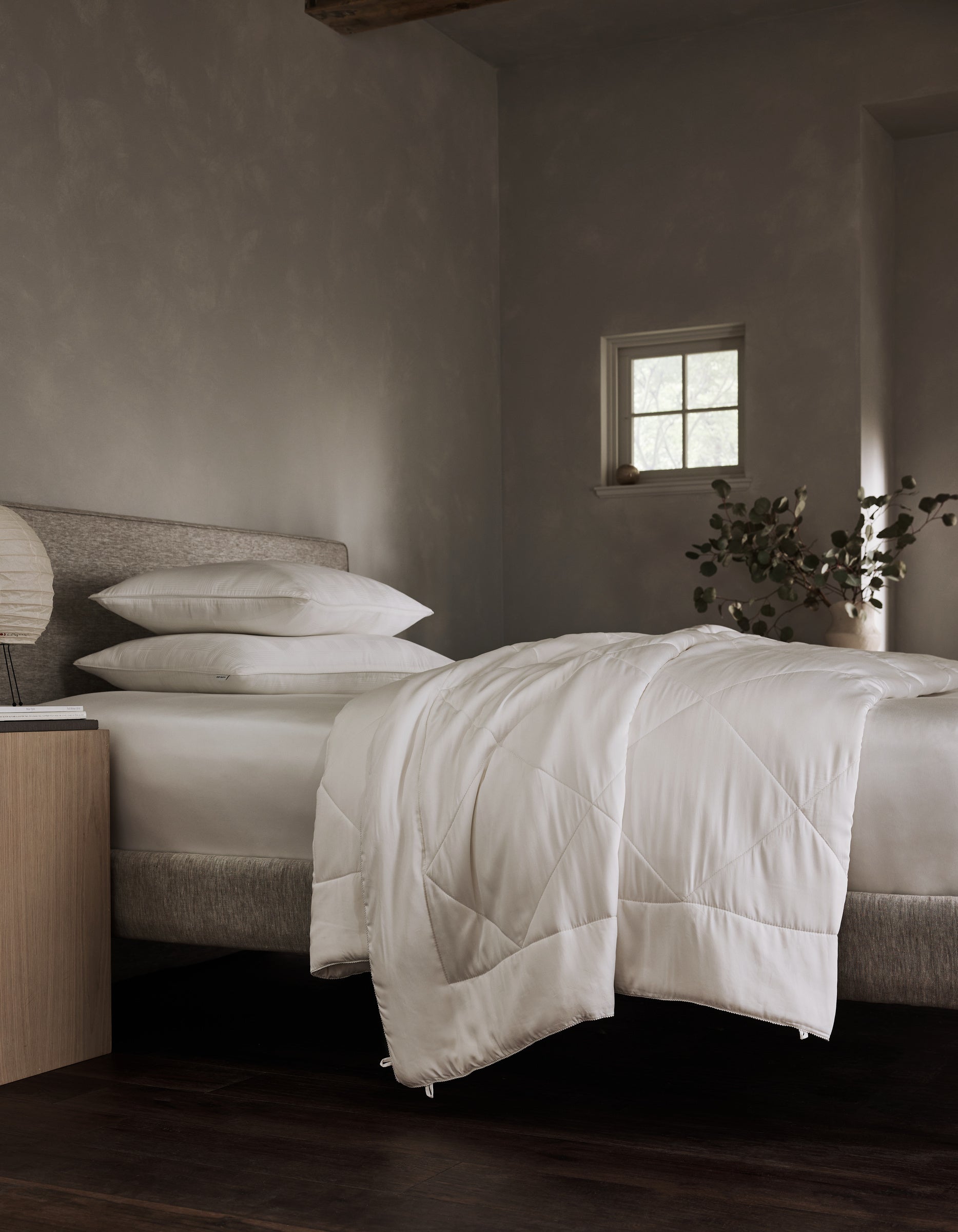 A neatly made bed features the Cozy Earth Bamboo Viscose Comforter with white bedding and pillows in a softly lit, minimalist room. A small window, bedside table, and leafy plant are seen in the background.|Filling:Standard 