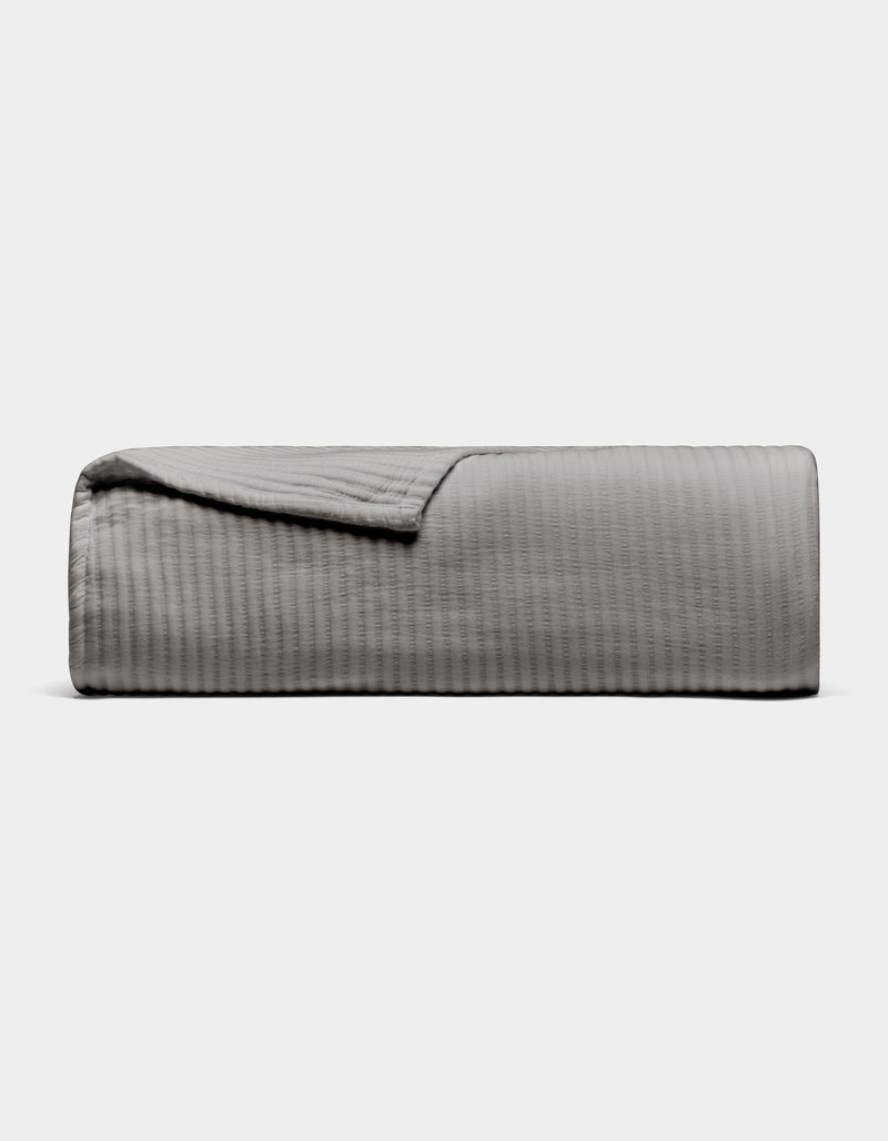 The Cozy Earth Bamboo Coverlet, shown neatly folded in light gray with a subtle textured pattern, is displayed on a plain white background. One corner is turned down to reveal the inner side of the coverlet.