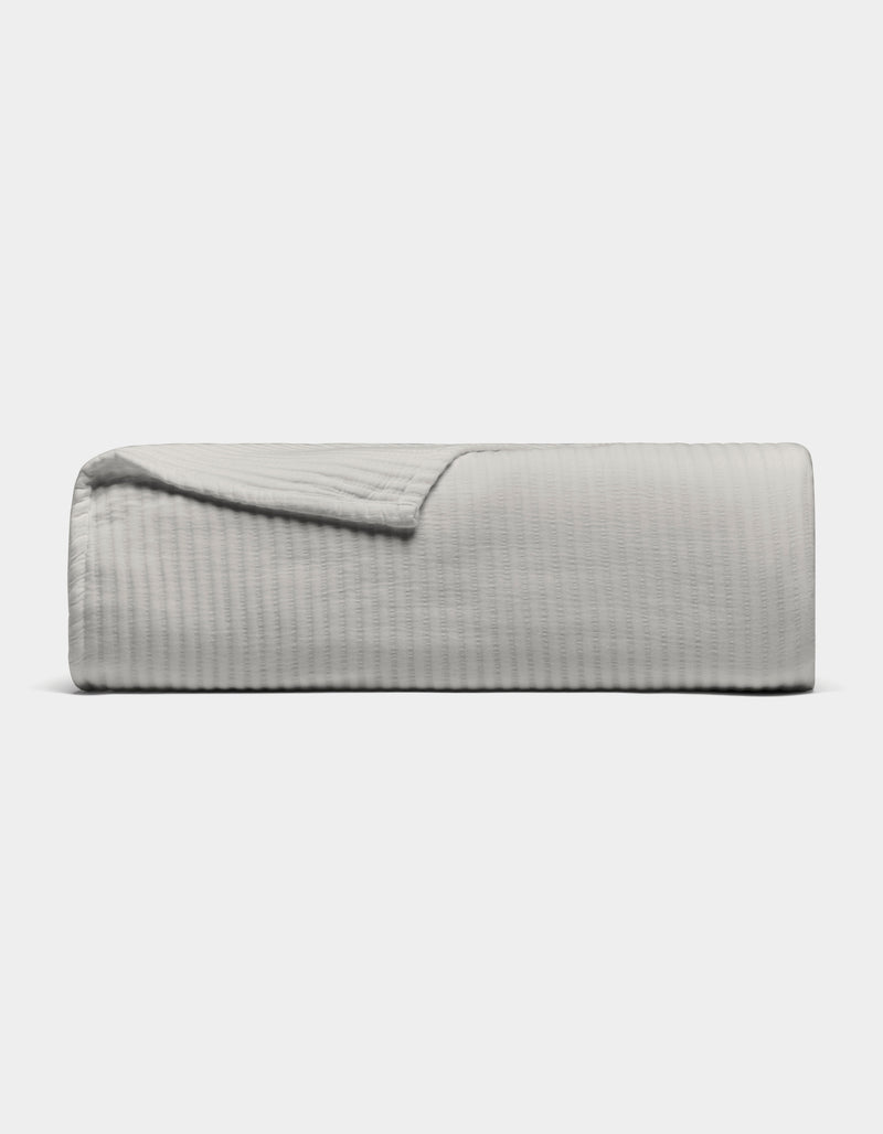 The Cozy Earth Bamboo Coverlet, shown in light gray with a textured design, is neatly folded against a plain white background. A corner is turned down to display its soft material.