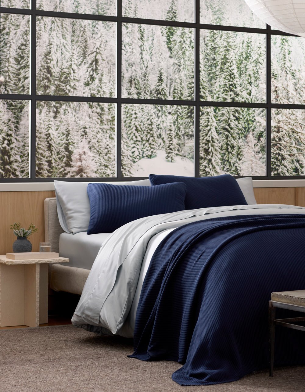 A neatly made bed featuring Cozy Earth's Bamboo Coverlet Shams in navy blue and crisp white sheets is beside a small nightstand. Large windows behind it showcase a snowy pine forest.