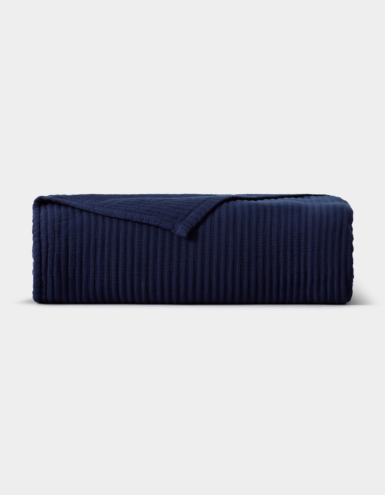 The Cozy Earth Bamboo Coverlet, in dark navy blue with a ribbed texture and one corner turned down, is displayed neatly against a plain light gray background.