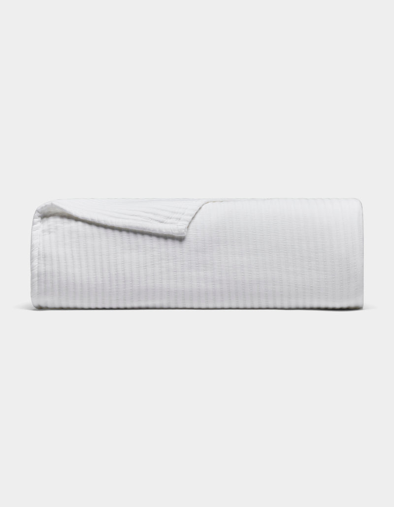 The Cozy Earth Bamboo Coverlet, a white textured blanket, is neatly folded with one corner slightly lifted, set against a plain light gray background.