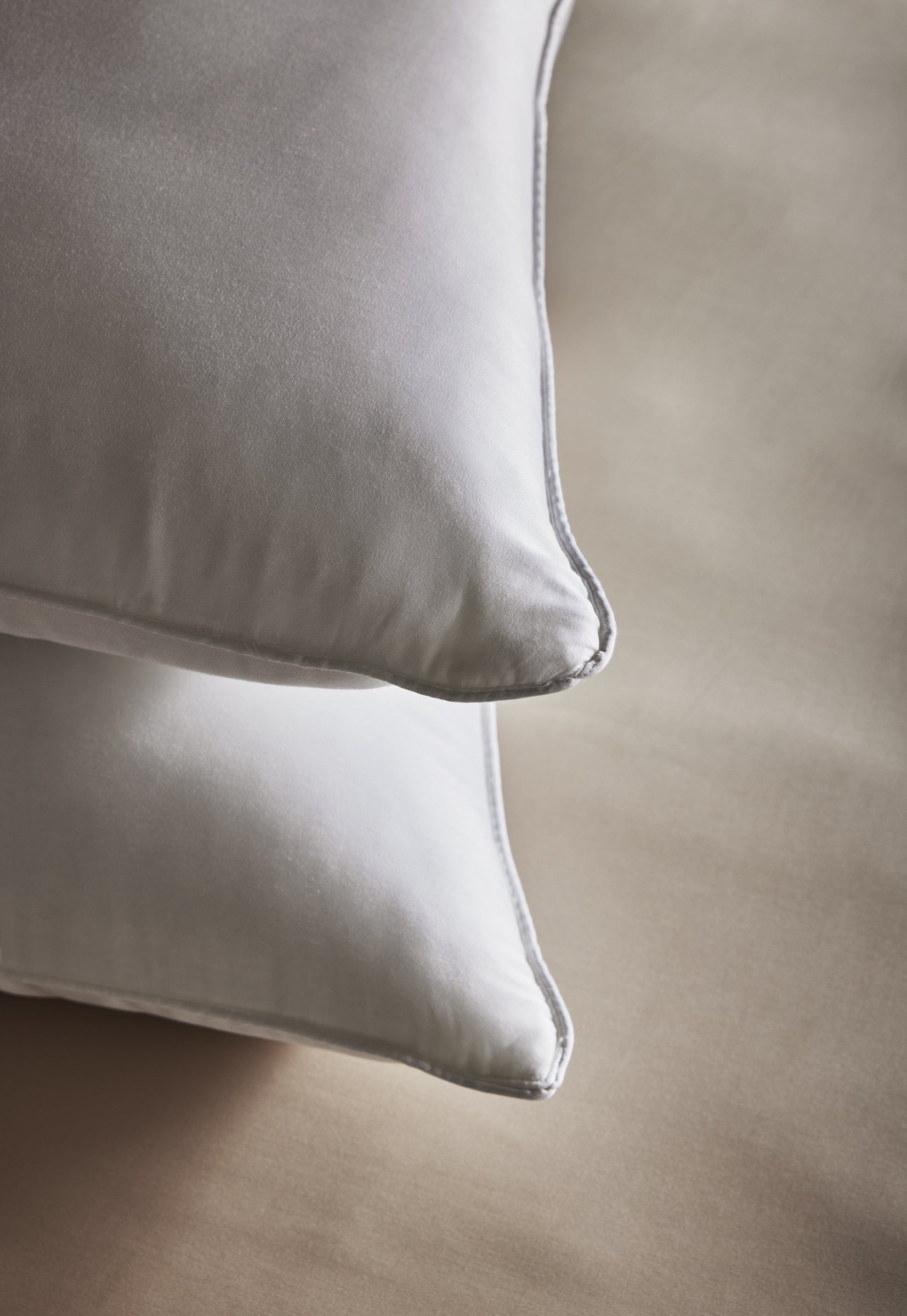 Two Bamboo Down Alternative Pillows by Cozy Earth with smooth, satiny pillowcases are neatly stacked on a light beige surface and viewed close up from the corner.