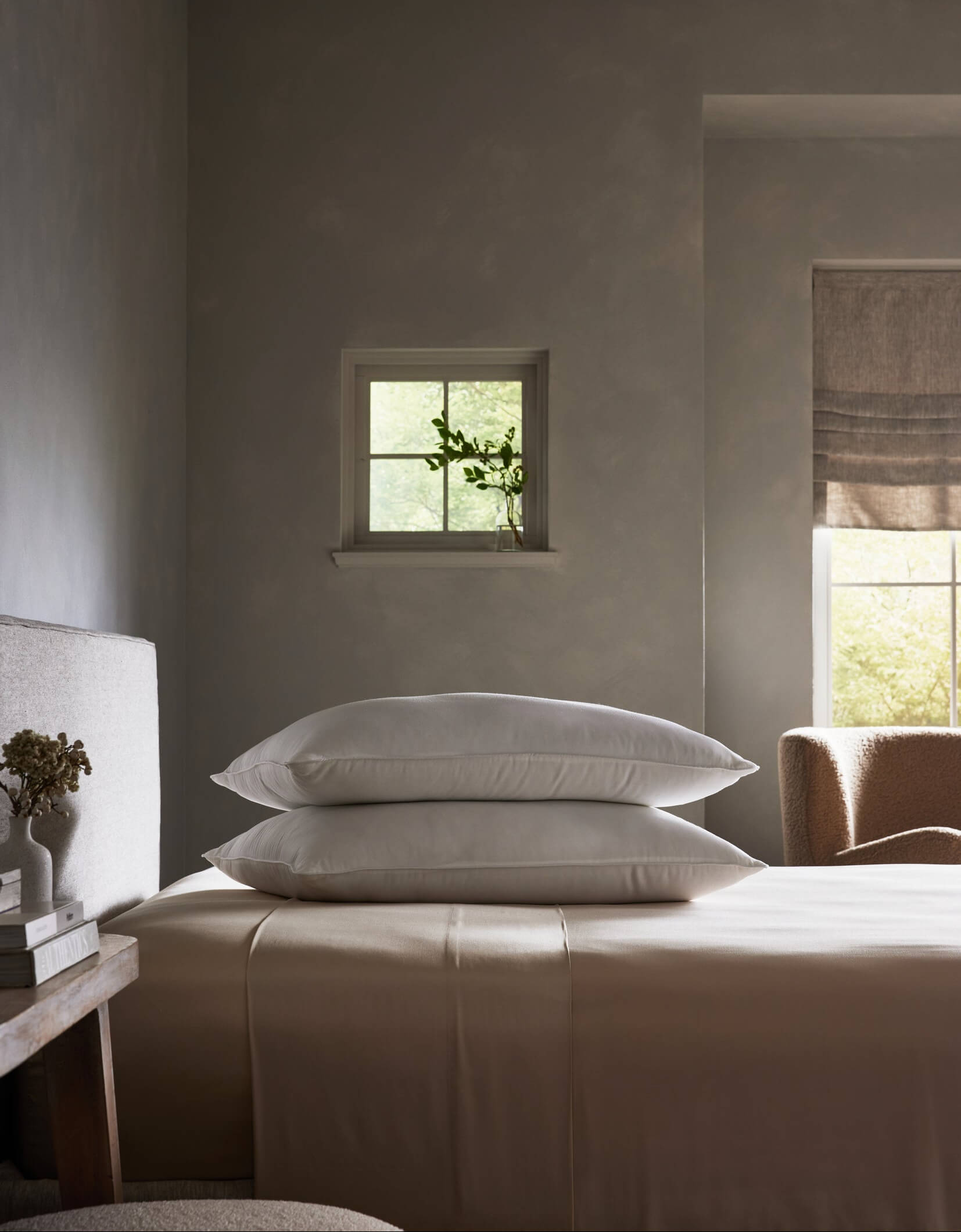 A serene, minimalist bedroom in neutral tones features a neatly made bed with the Cozy Earth Down Alternative Insert Bundle and two stacked white pillows, softly illuminated by daylight near a window with a glass vase of greenery.