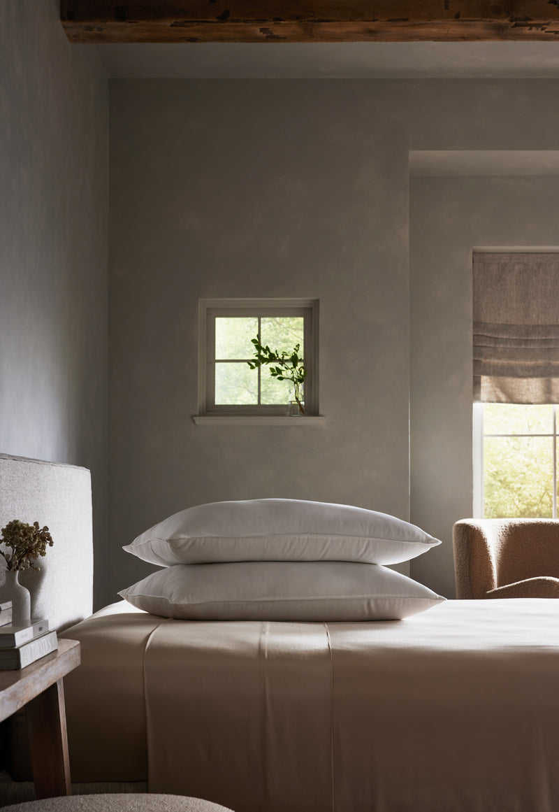 A serene bedroom with neutral tones features a neatly made bed topped with Cozy Earth Bamboo Down Alternative Pillows, a small window with a potted plant, and soft natural light streaming in from a larger window with a beige shade.Standard King