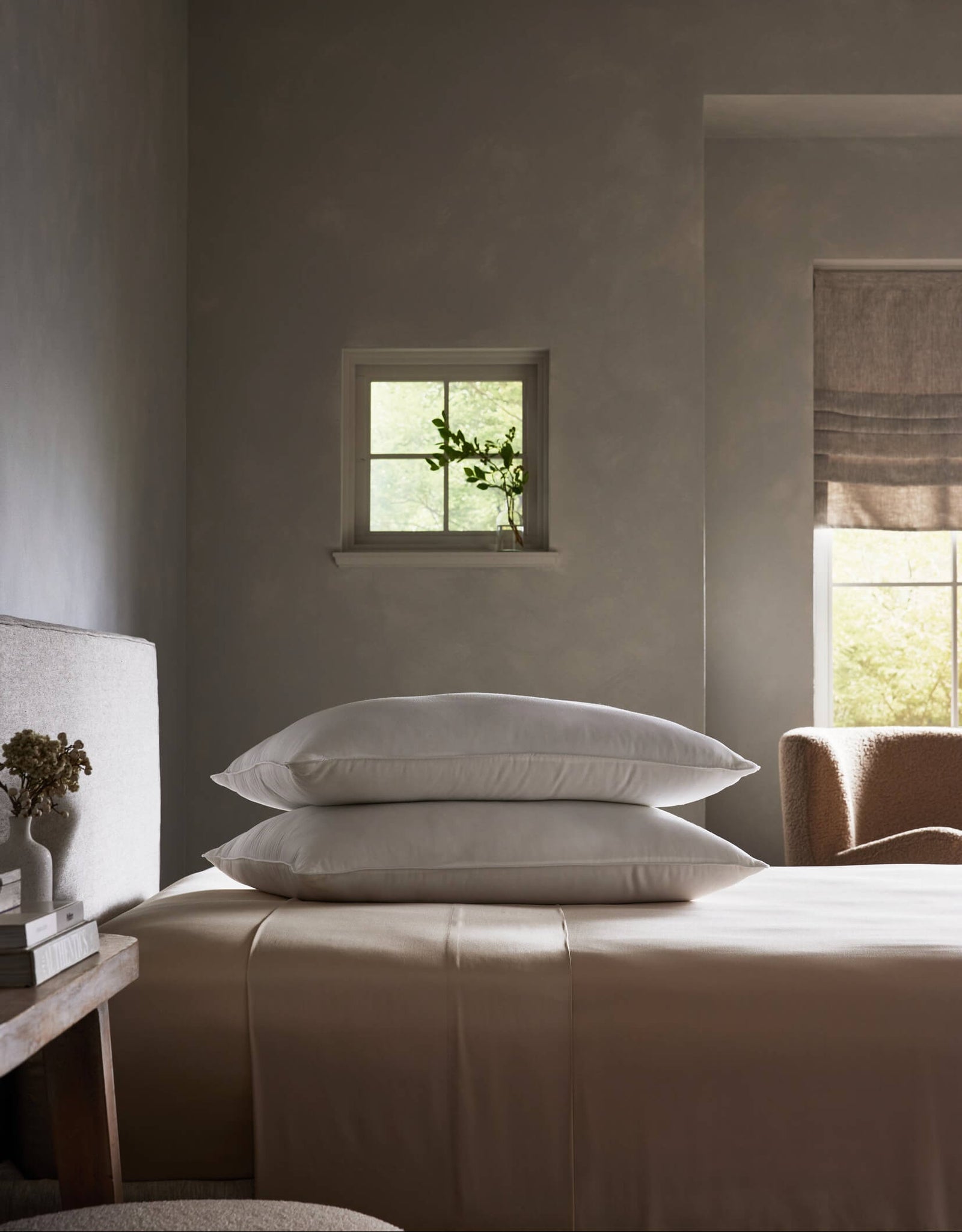 A neatly made bed features Cozy Earth’s Down Alternative Insert Bundle with two stacked white pillows in a minimalist, softly lit bedroom. A small window with a vase plant and another window with a beige shade create a serene mood.
