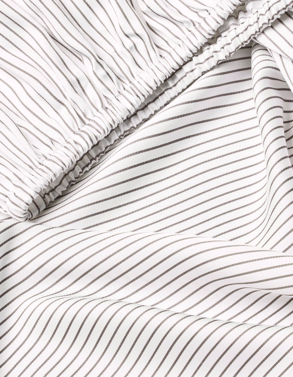 A close-up of Cozy Earth’s Bamboo Fitted Sheet shows soft white fabric with thin black pinstripes, light wrinkles, and an elastic band along the edge.