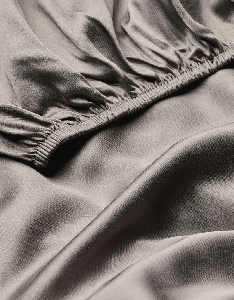 A close-up of the Cozy Earth Bamboo Fitted Sheet shows smooth, light gray satin fabric with gentle folds and a soft, shiny surface. An elastic gathered seam runs diagonally across the image.