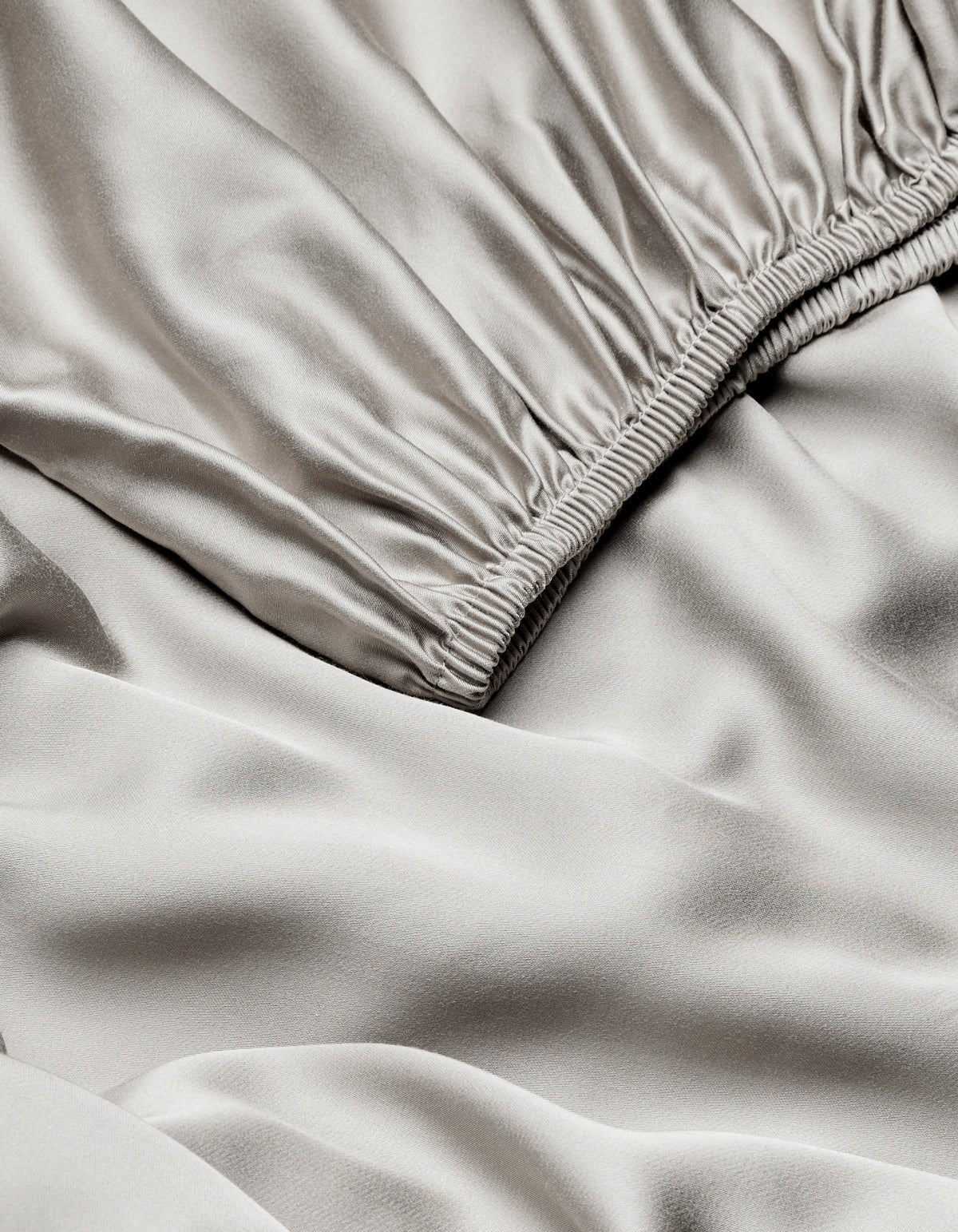Close-up of Cozy Earth Bamboo Fitted Sheet in light gray, featuring a gathered elastic waistband and showcasing its smooth, shiny, slightly wrinkled satin texture.|Color:Light Grey