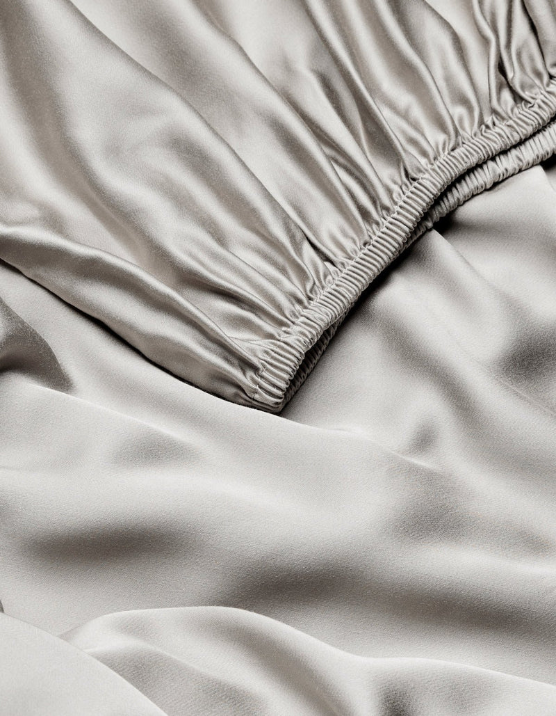 Close-up of Cozy Earth Bamboo Fitted Sheet in light gray, featuring a gathered elastic waistband and showcasing its smooth, shiny, slightly wrinkled satin texture.