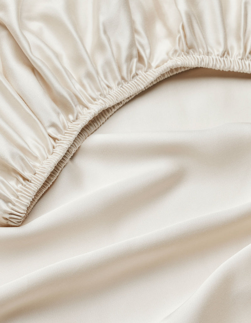 Close-up of Cozy Earth's Bamboo Fitted Sheet in smooth, ivory-colored satin with an elastic edge, highlighting its shiny, soft texture.