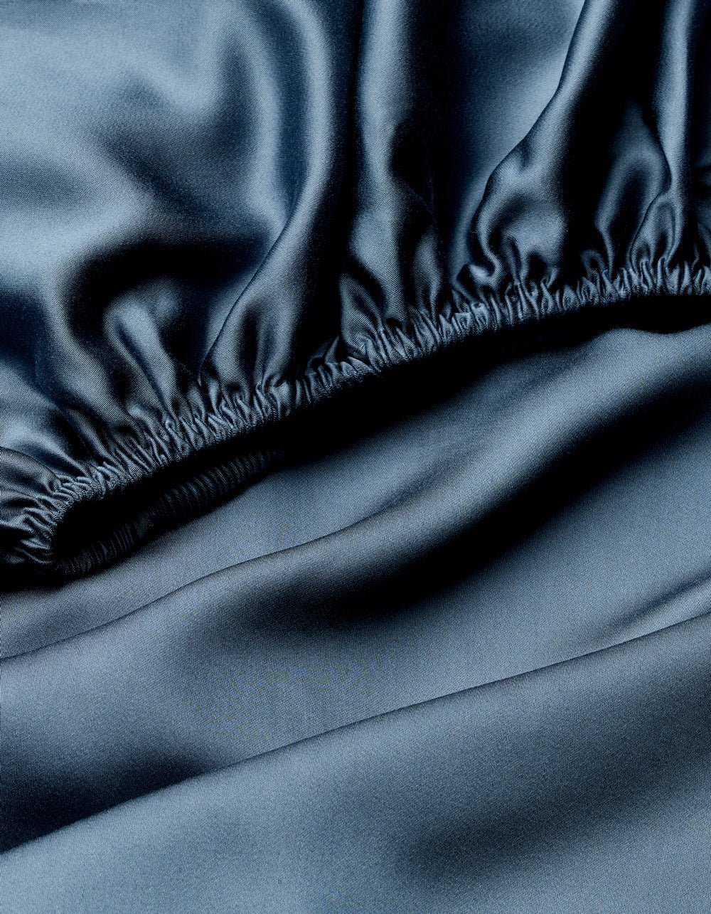 A close-up of dark, shiny fabric with a silky texture and reflective sheen shows the gathered, elastic edge of the Cozy Earth Bamboo Fitted Sheet.|Color:Pacific Blue