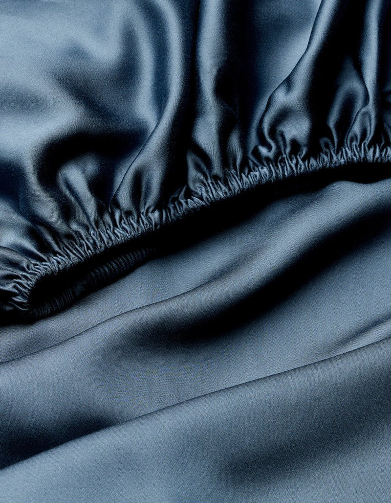A close-up of dark, shiny fabric with a silky texture and reflective sheen shows the gathered, elastic edge of the Cozy Earth Bamboo Fitted Sheet.