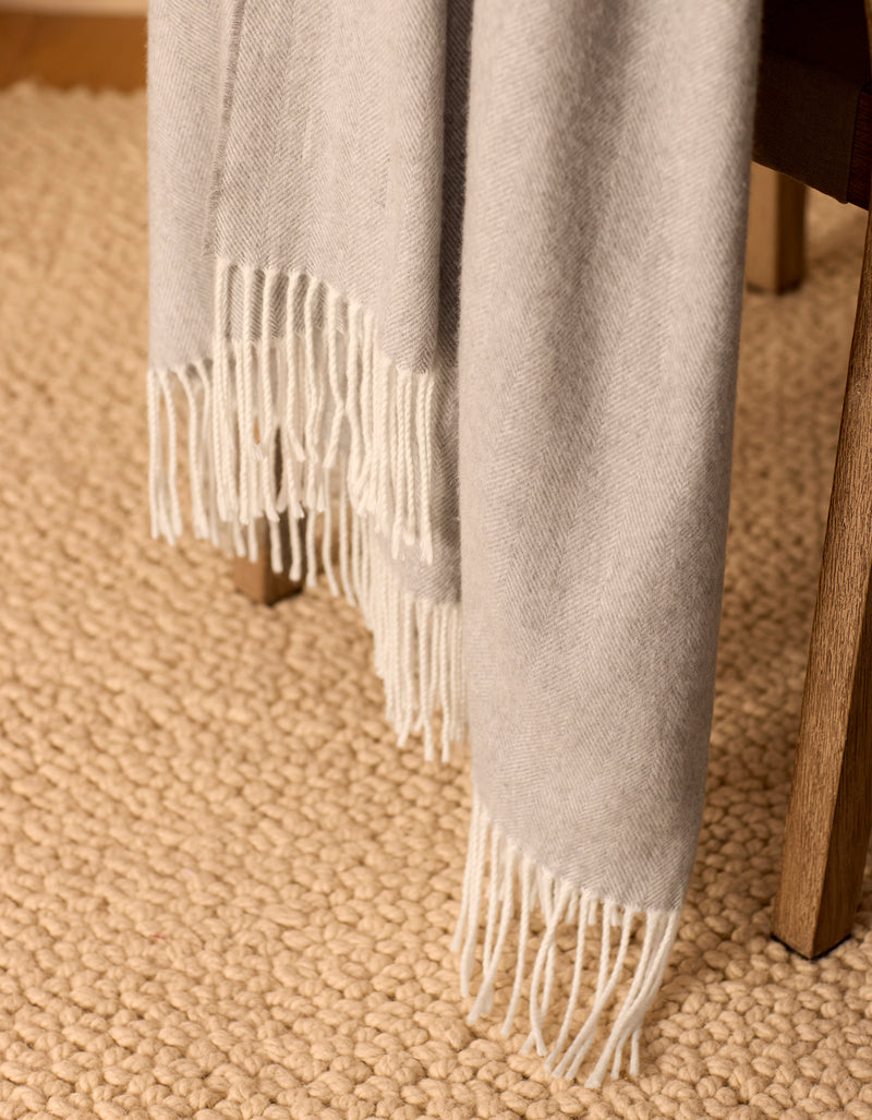 The Bamboo Herringbone Tassel Throw by Cozy Earth, in light gray, is draped over a wooden chair with a textured beige rug placed on the floor beneath.