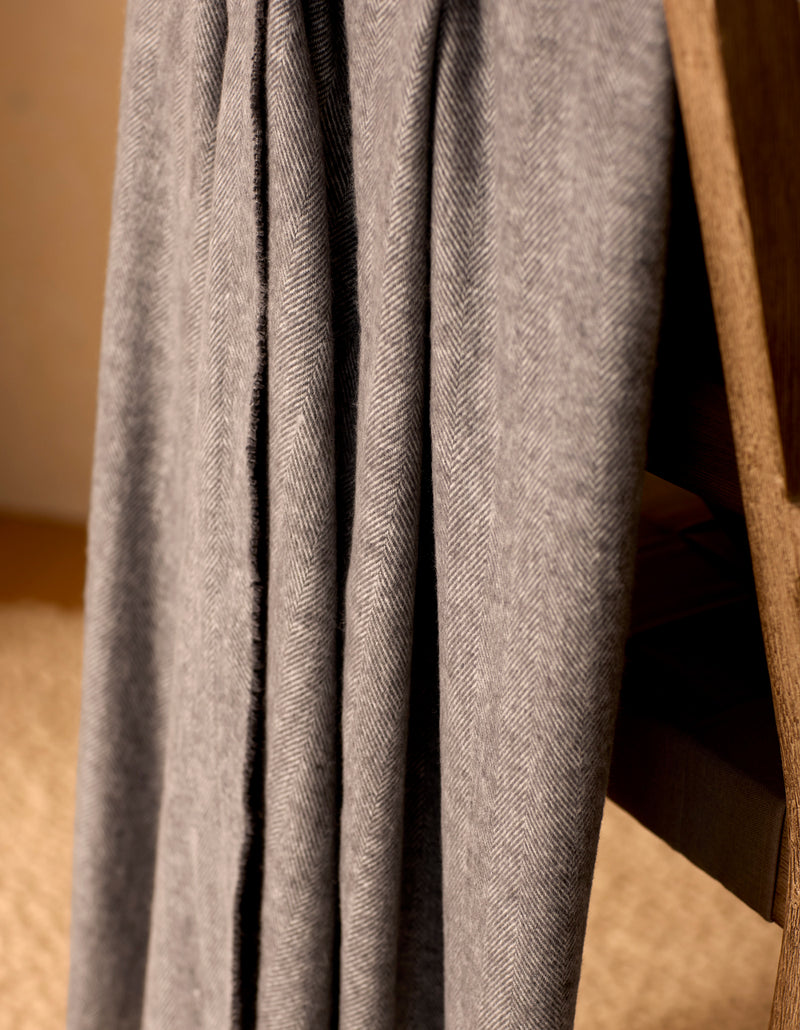A close-up of the Cozy Earth Bamboo Herringbone Tassel Throw, featuring a gray herringbone pattern, draped over a wooden chair against a blurred beige background.