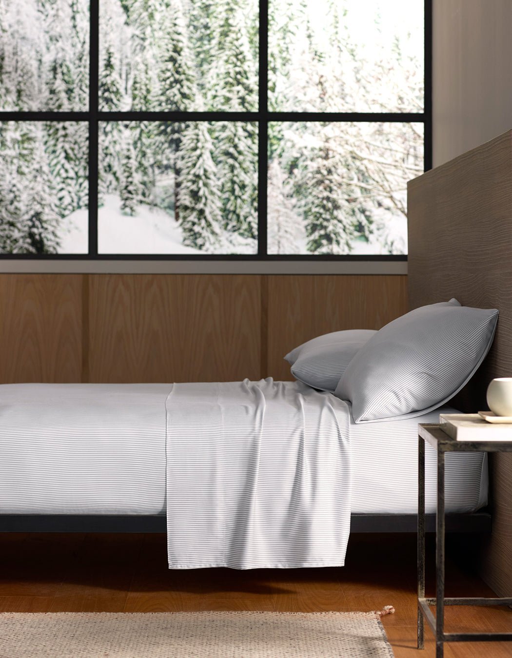 A bed with Cozy Earth Bamboo Flat Sheets and pillows is beside a small table with a cup. Large windows reveal a snowy forest, filling the room with natural light.