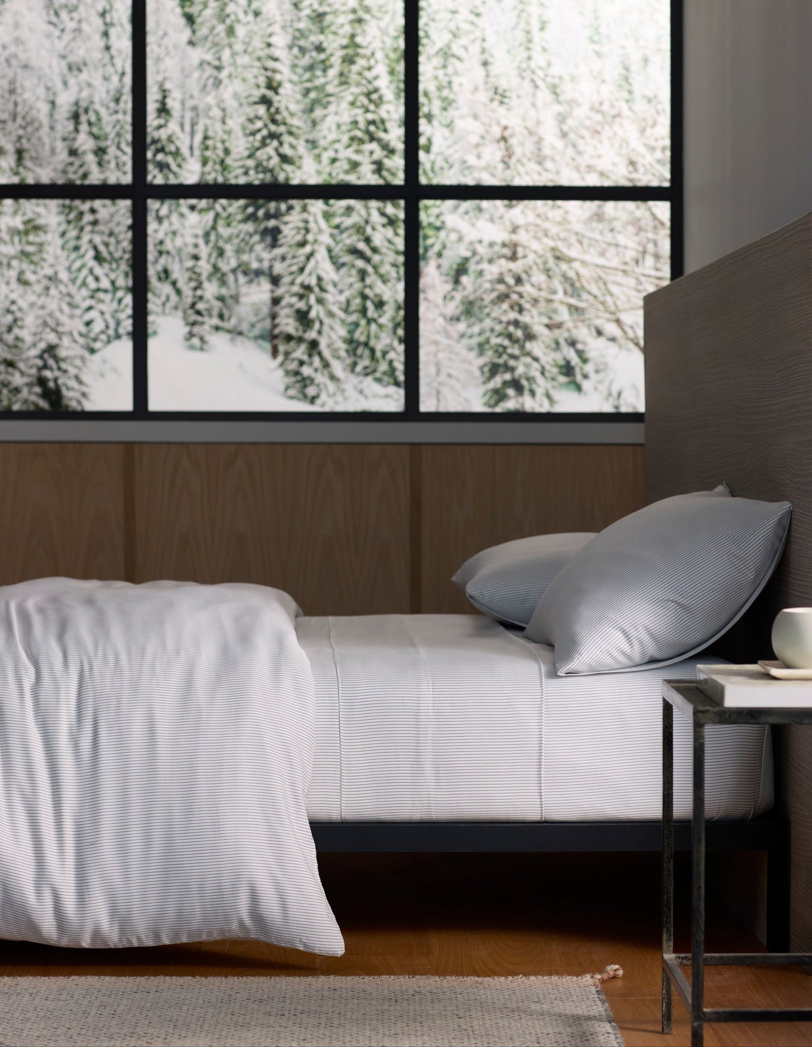 A neatly made bed with Cozy Earth’s Bamboo Sheet Set stands next to a small table with a cup, set against large windows overlooking a snowy forest scene. 