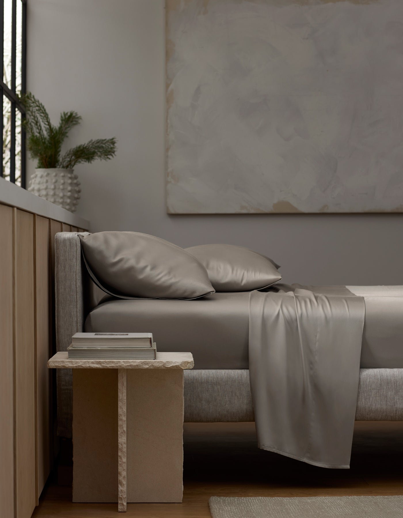 A modern, minimal bedroom features a gray upholstered bed dressed in Cozy Earth’s Bamboo Sheet Set and matching pillows. A stone nightstand holds books and a vase with greenery, while large abstract art hangs on the neutral wall.