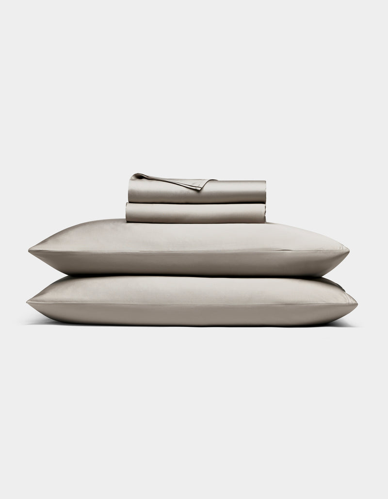 The Cozy Earth Bamboo Sheet Set, in light gray, includes neatly folded bed sheets and pillowcases stacked atop two pillows, all shown against a plain white background.