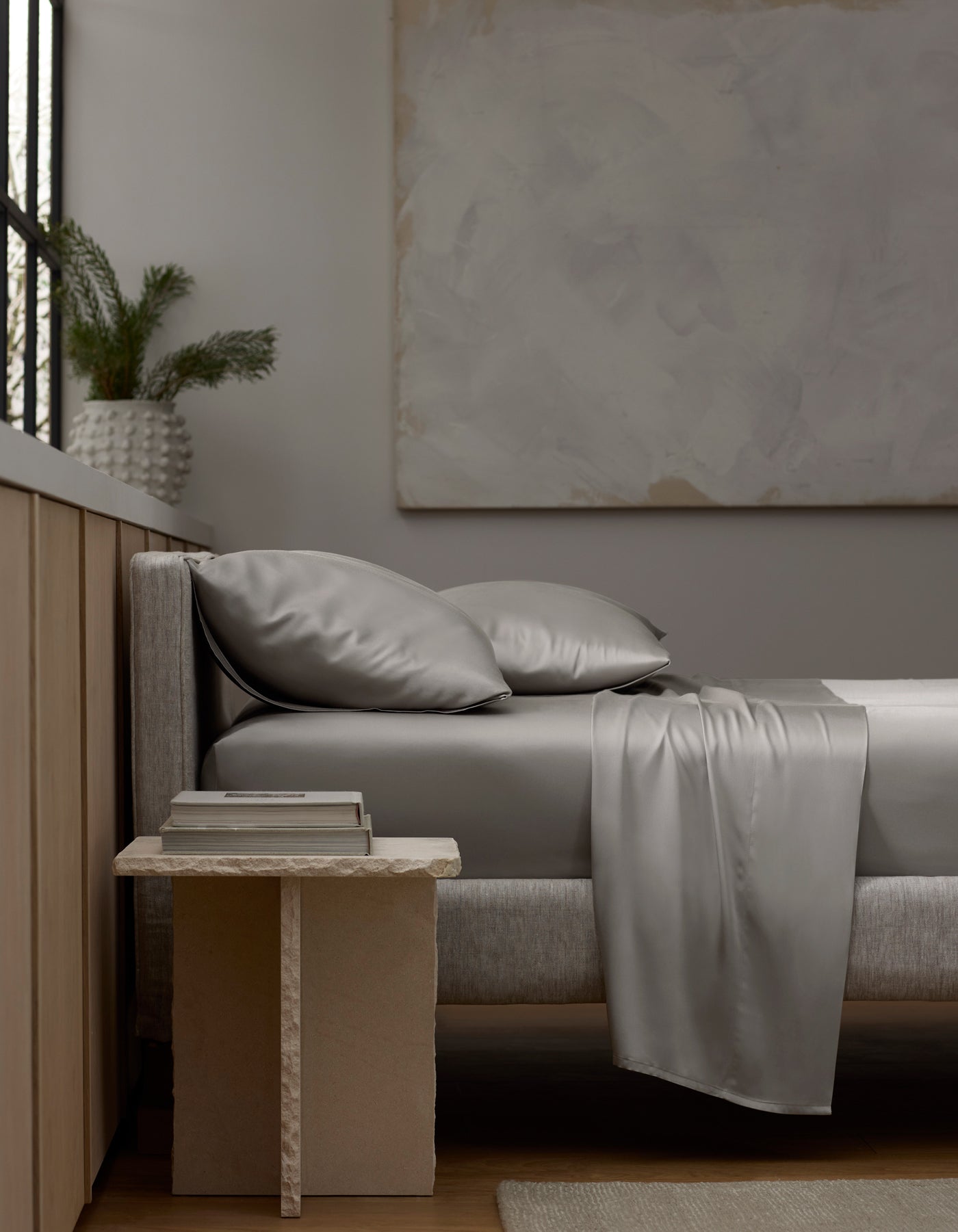 A modern bedroom with a neatly made bed featuring Cozy Earth’s Bamboo Sheet Set in shiny silver-gray. A textured side table holds stacked books, and a vase with greenery sits on the windowsill, creating minimalist neutral-toned decor. 