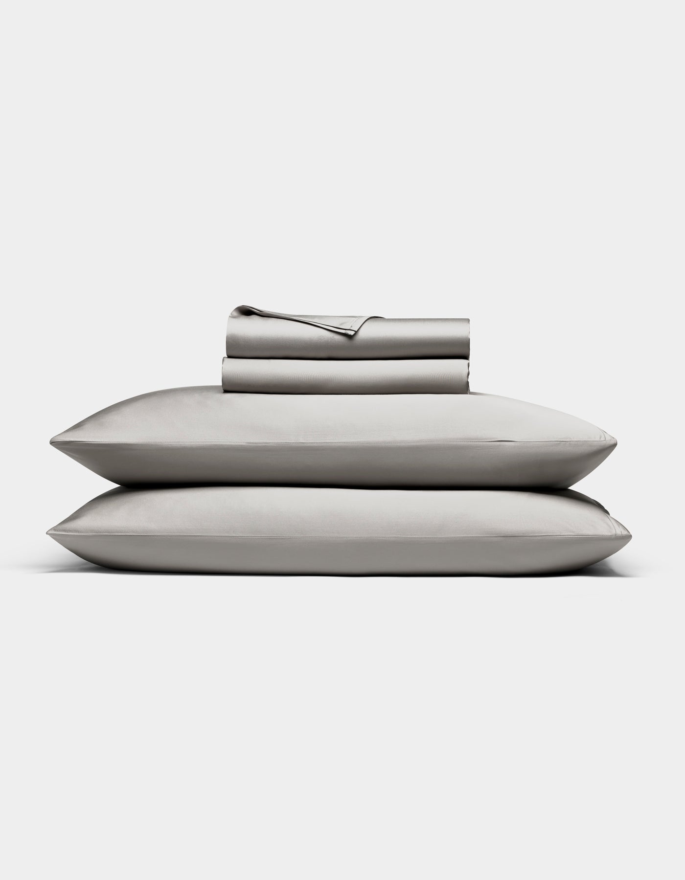 The Bamboo Sheet Set by Cozy Earth in light gray is neatly folded, with two pillows stacked and both flat and fitted sheets rolled on top, all displayed against a white background.