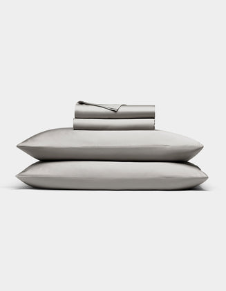 The Bamboo Sheet Set by Cozy Earth in light gray is neatly folded, with two pillows stacked and both flat and fitted sheets rolled on top, all displayed against a white background.|Color:Light Grey