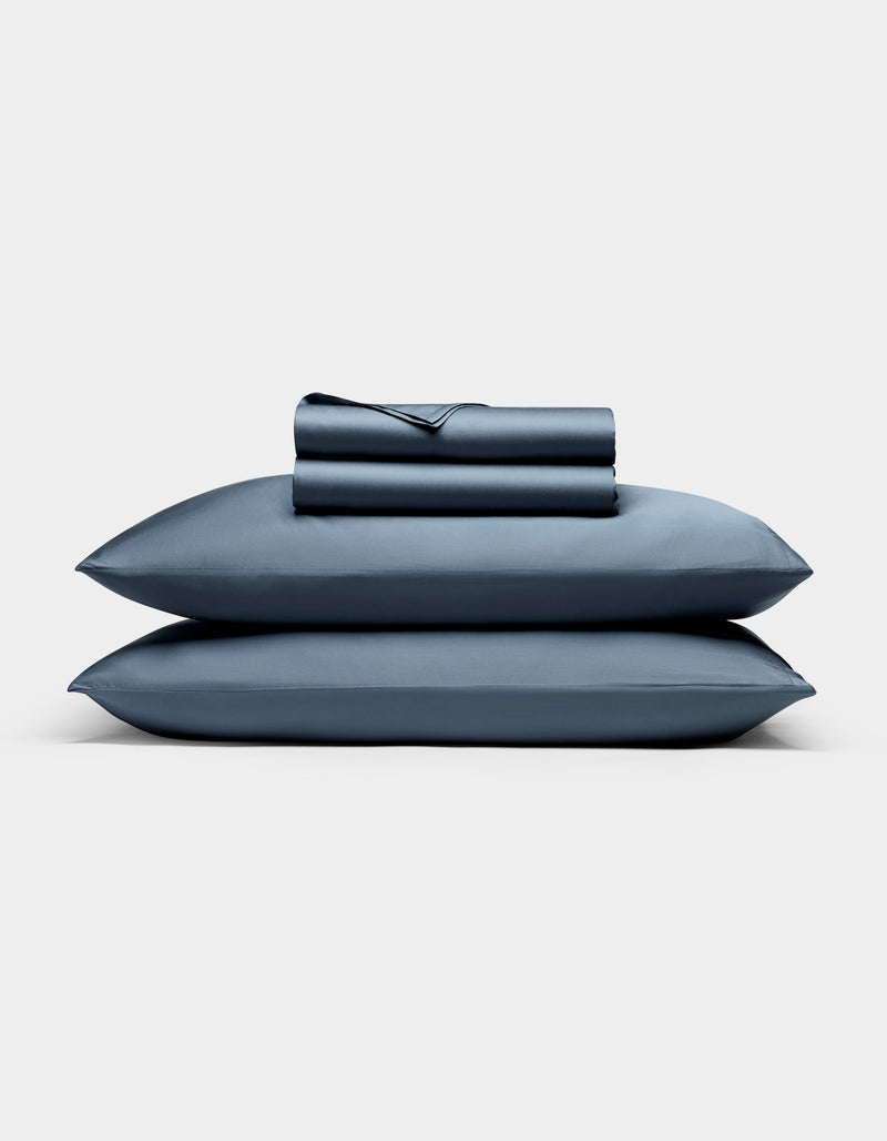 A neatly folded Cozy Earth Bamboo Sheet Set, featuring a fitted sheet, flat sheet, and two pillowcases, is stacked atop two matching blue pillows against a plain white background.