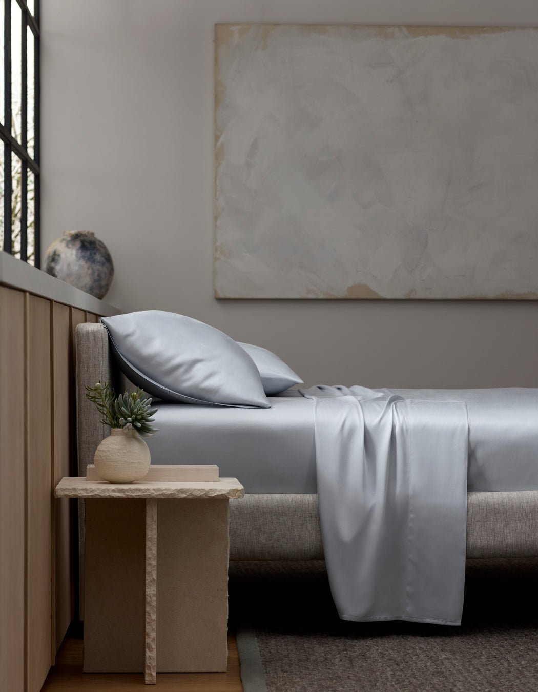 A minimalist bedroom with a light gray upholstered bed, Cozy Earth Bamboo Sheet Set in silver-gray, two pillows, a beige nightstand with a small potted plant, a large abstract painting, and a window letting in natural light.