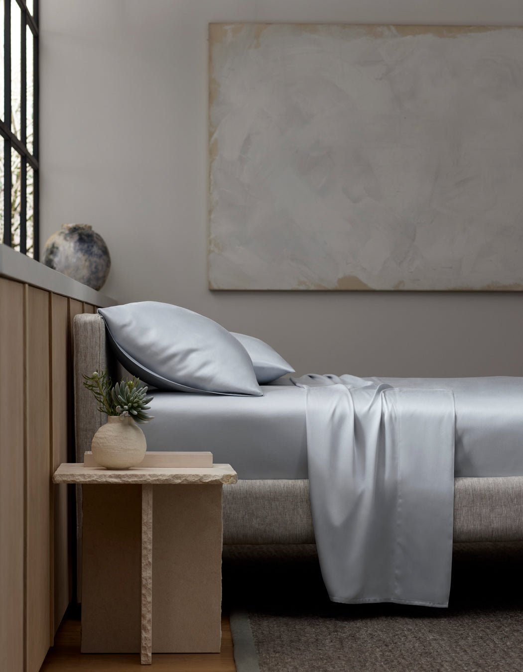 A minimalist bedroom with a light gray upholstered bed, Cozy Earth Bamboo Sheet Set in silver-gray, two pillows, a beige nightstand with a small potted plant, a large abstract painting, and a window letting in natural light.