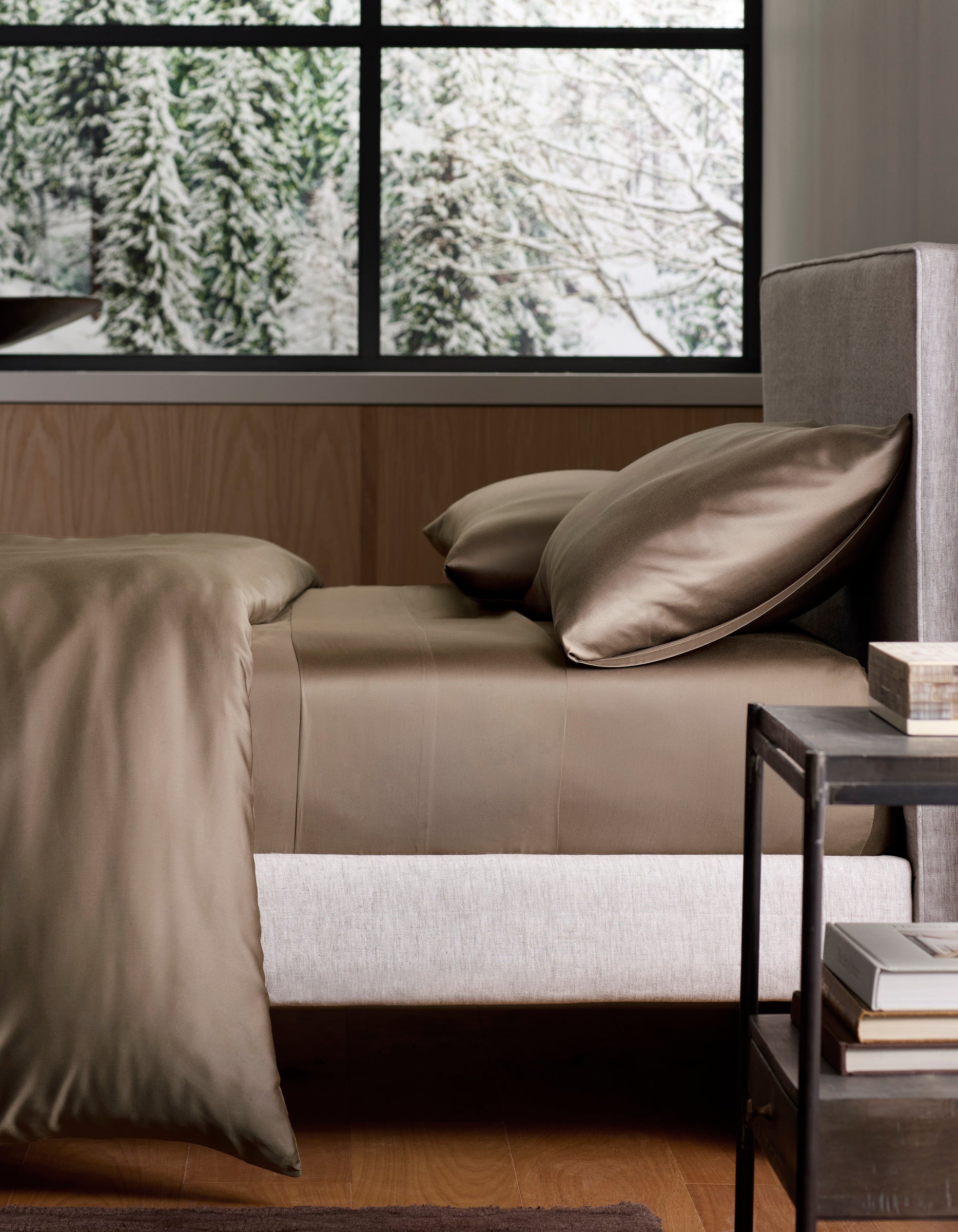A neatly made bed with Cozy Earth Bamboo Sheet Set in silky brown lies next to a black nightstand, framed by a large window revealing snow-covered trees outside. 