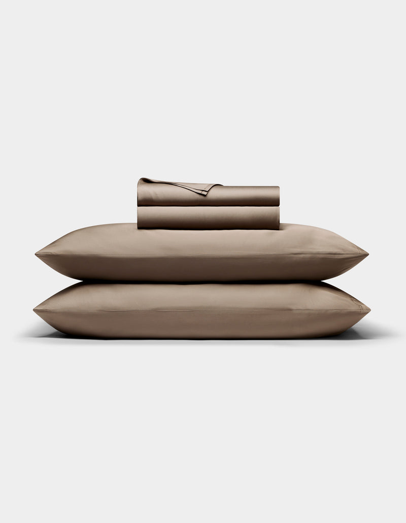 A neatly folded Cozy Earth Bamboo Sheet Set in taupe, with pillowcases, is stacked atop two matching pillows against a plain white background.