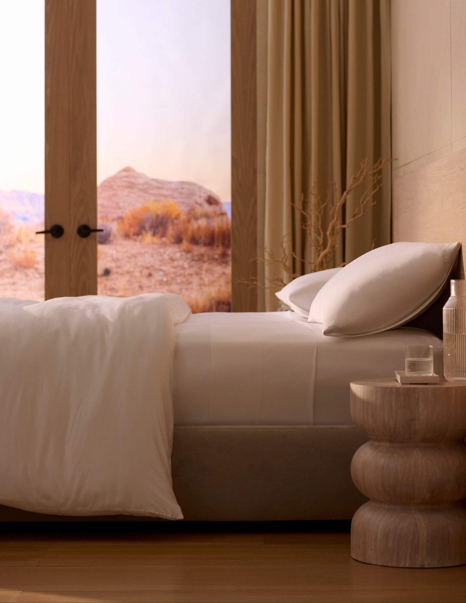 A bed made with Cozy Earth's Bamboo Sheet Set features crisp white linens beside a wooden nightstand with a glass of water and bottle. Large windows reveal a scenic desert landscape, while neutral tones create a calm, minimalist vibe. 