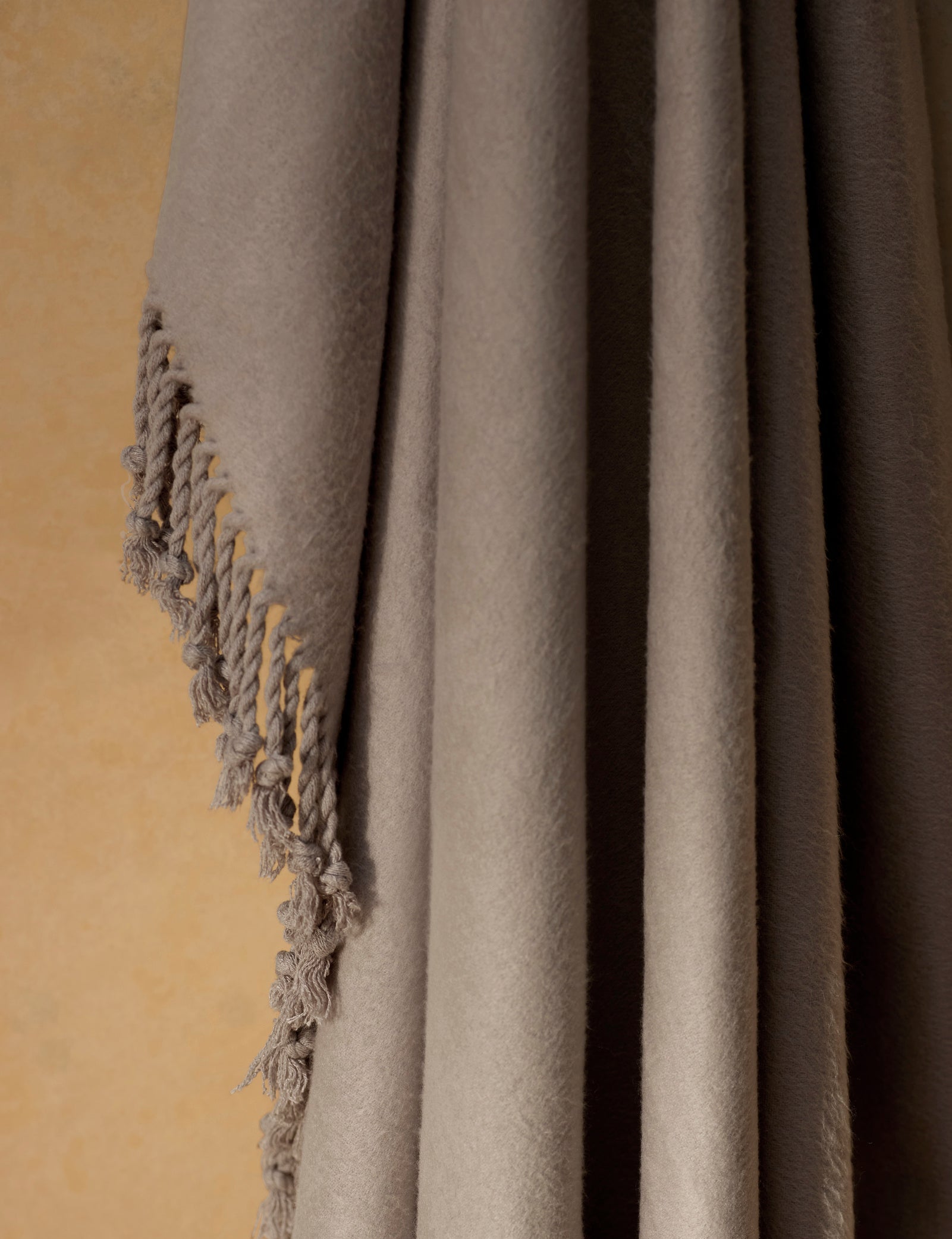 The Cozy Earth Bamboo Tassel Throw, in soft light brown, hangs against a beige wall and features a fringe detail with twisted threads along one edge.