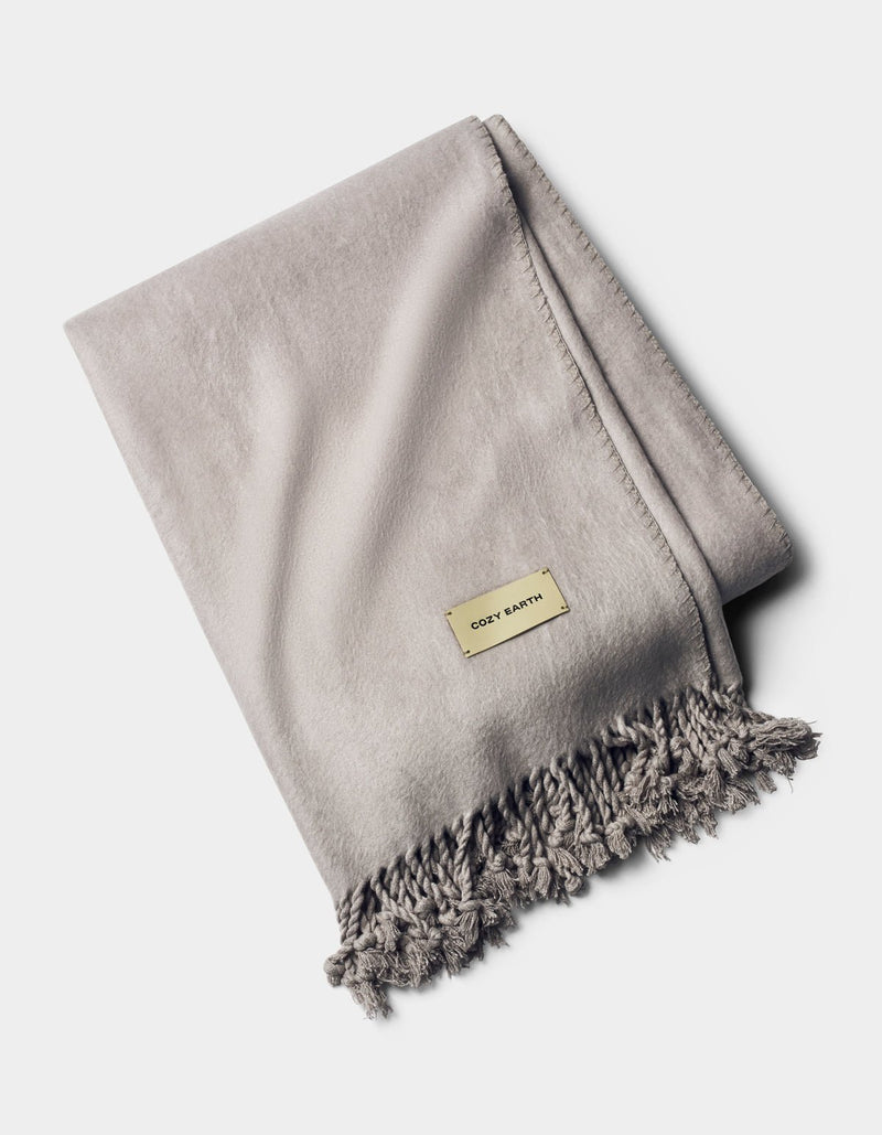 A neatly folded light gray Bamboo Tassel Throw with fringe on one edge and a small rectangular Cozy Earth label on top, displayed against a plain white background.