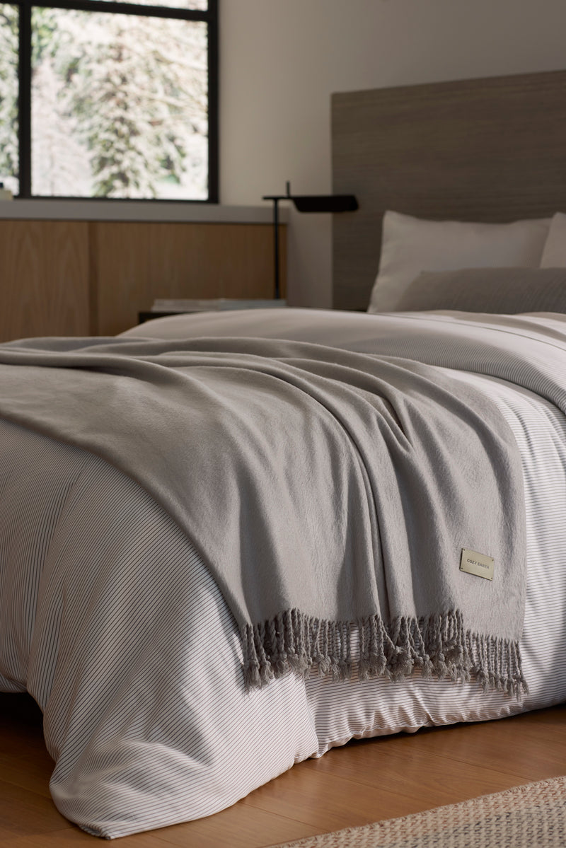 A neatly made bed with white and light gray bedding is topped with the Cozy Earth Bamboo Tassel Throw in gray. The room features a wooden headboard, light wood furniture, and a window letting in natural light.