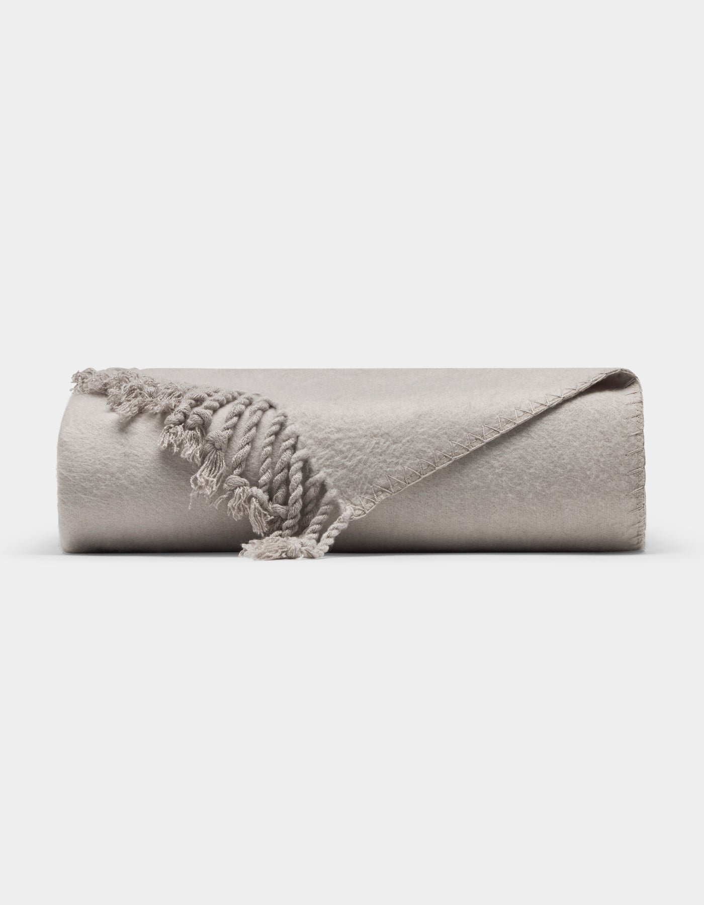 The Cozy Earth Bamboo Tassel Throw is a neatly rolled, light gray blanket with fringed tassels, shown on a white background. One corner is flipped to reveal its soft texture and detailed edging.|Color:Dove Grey