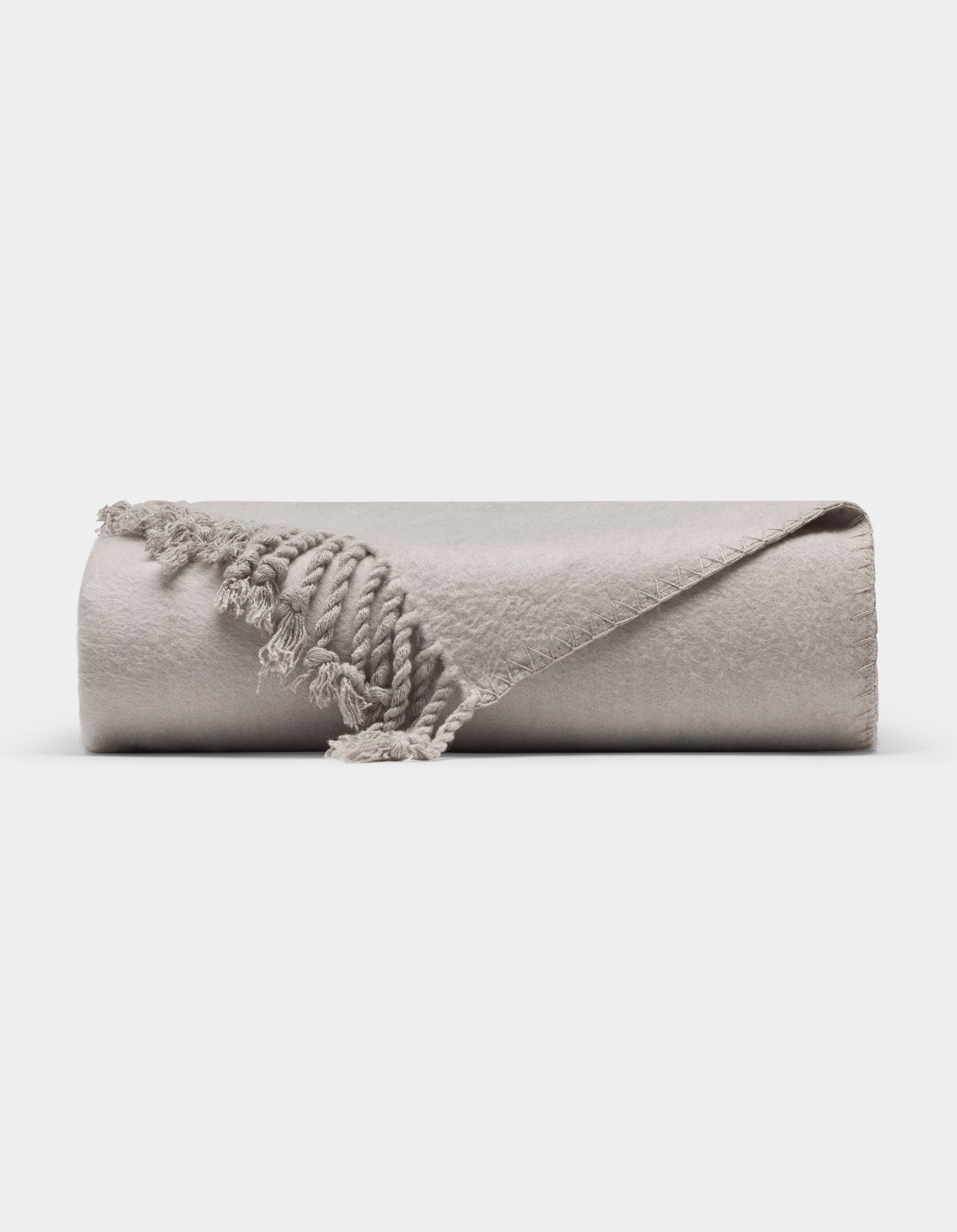 The Cozy Earth Bamboo Tassel Throw is a neatly rolled, light gray blanket with fringed tassels, shown on a white background. One corner is flipped to reveal its soft texture and detailed edging.|Color:Dove Grey
