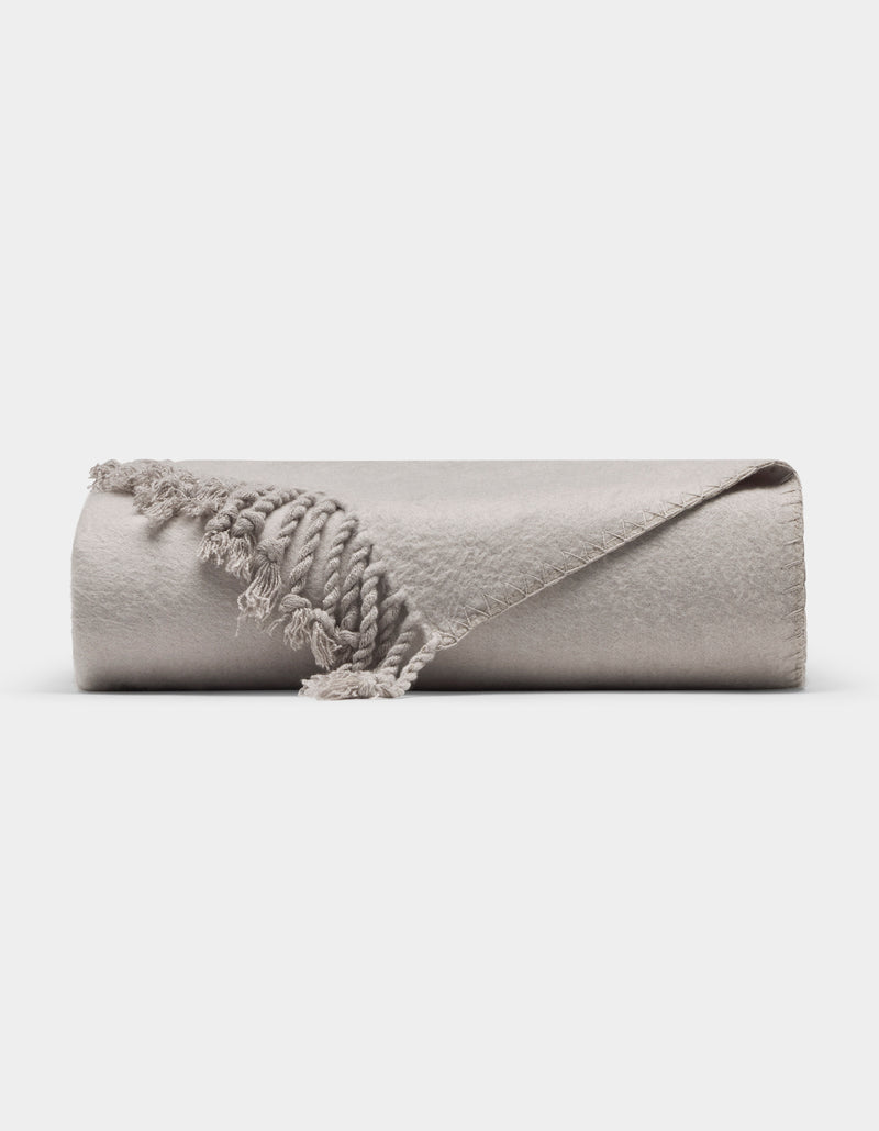 The Cozy Earth Bamboo Tassel Throw is a neatly rolled, light gray blanket with fringed tassels, shown on a white background. One corner is flipped to reveal its soft texture and detailed edging.