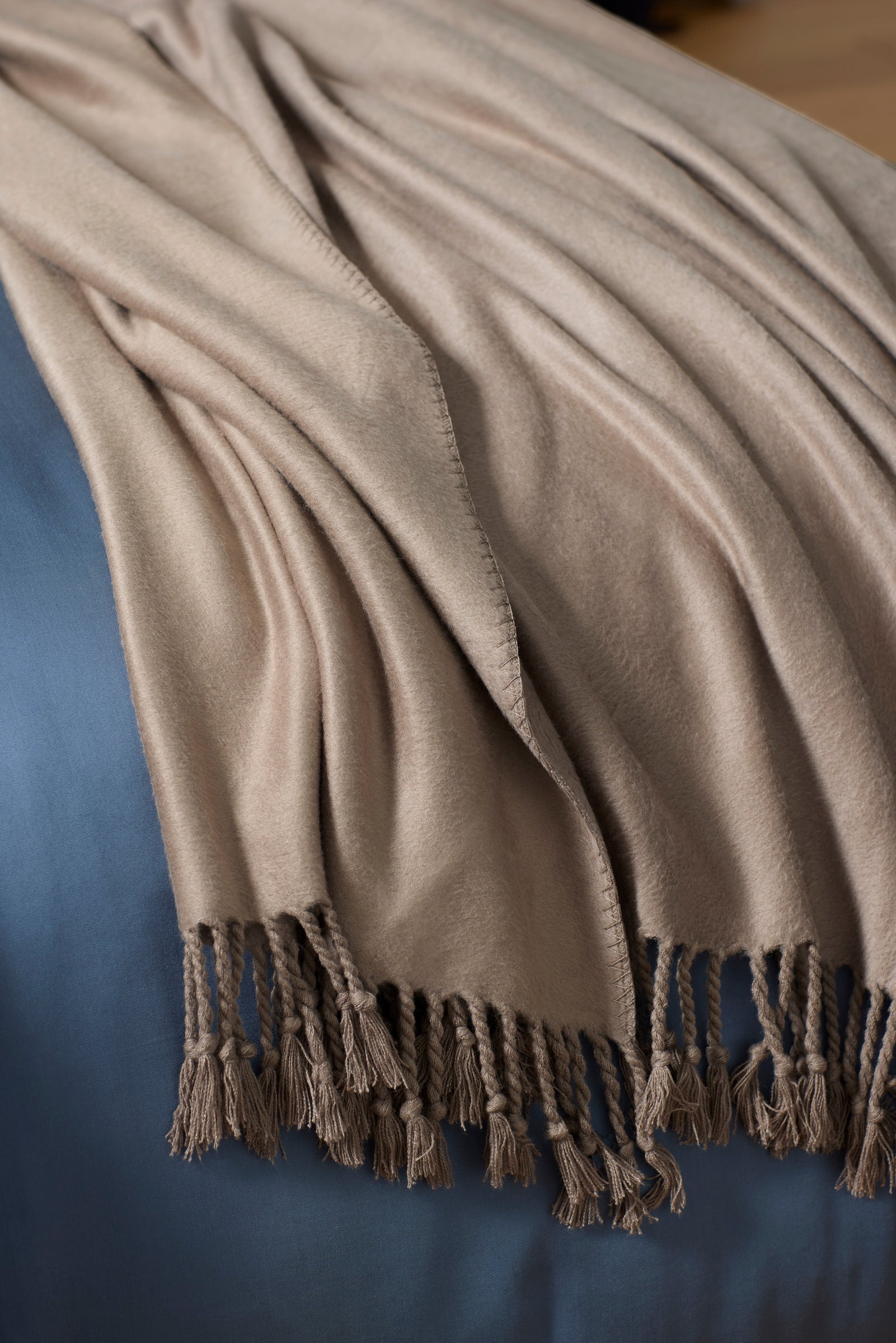 The Cozy Earth Bamboo Tassel Throw, featuring tasseled edges, is draped over a blue surface, forming soft folds and a cozy look.|Color:Walnut