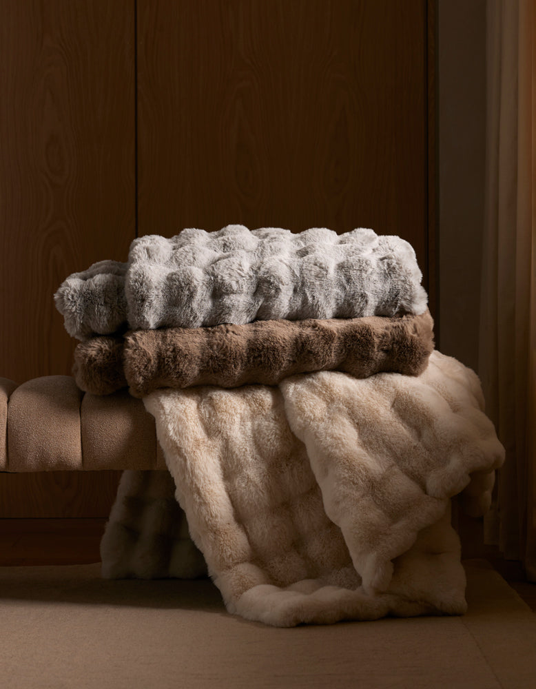 Stack of textured Bubble Cuddle pillows and Bubble Cuddle blankets on a padded bench against a wooden wall.