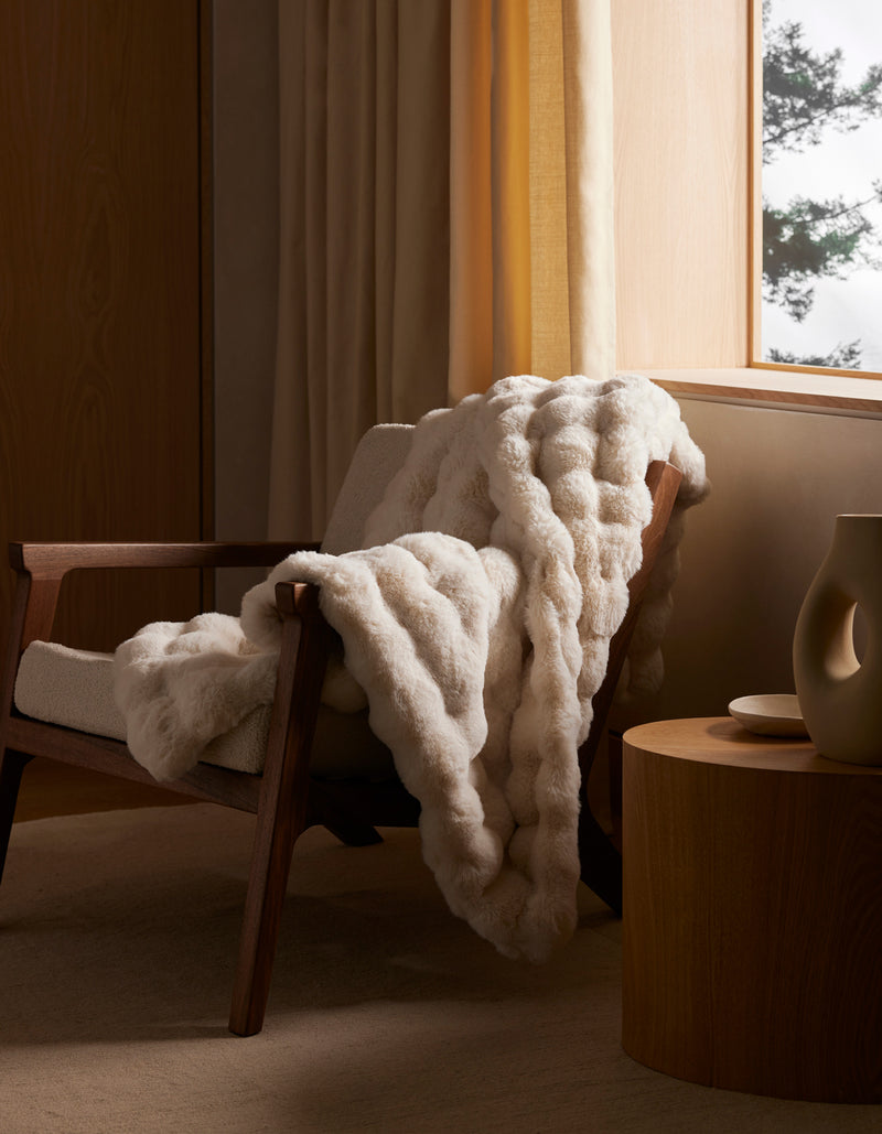 The Cozy Earth Bubble Cuddle Blanket, in cream, is draped over a wooden armchair next to a round wooden side table with beige curtains and soft natural light filtering through the window.